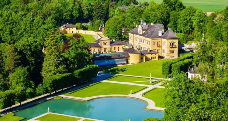 Un palacio barroco amarillo brillante rodeado de jardines cuidados, piscinas y frondoso bosque