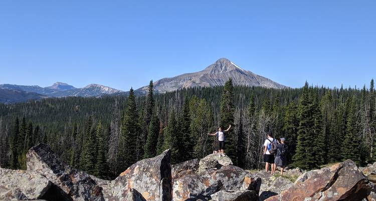 Los excursionistas están de pie en una saliente rocosa con los brazos levantados hacia las montañas lejanas cubiertas de coníferas.