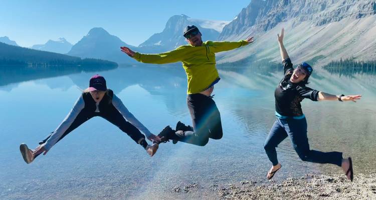 Tres amigos saltan alegremente junto a un lago alpino tranquilo como un espejo que refleja picos majestuosos.