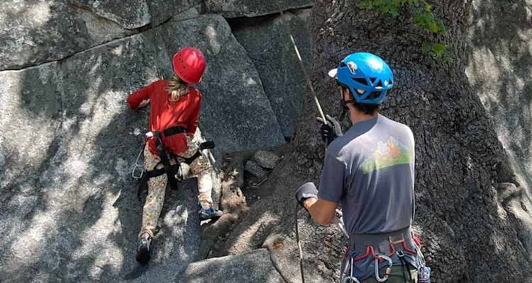 L'instructeur assure un jeune grimpeur qui monte une dalle de granit à l'ombre.