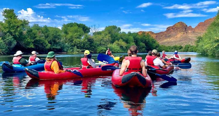 Une flottille de kayaks rouges et bleus avec leurs pagayeurs dérive sur une rivière calme encadrée par des arbres verts et des collines désertiques.