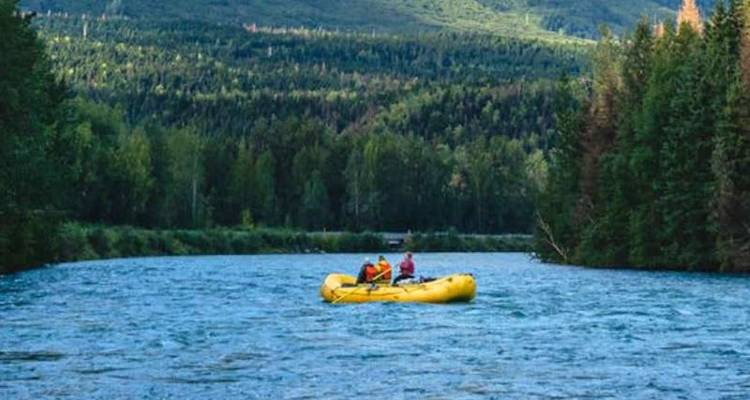 Deux personnes font descendre un petit radeau jaune sur une large rivière bleue bordée par une forêt dense de conifères.