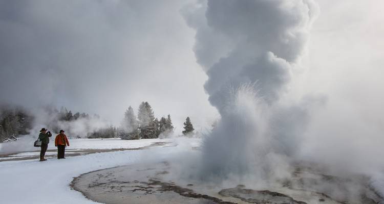 Un geyser entre en éruption au milieu d'un paysage enneigé tandis que deux visiteurs bien couverts observent depuis la passerelle.