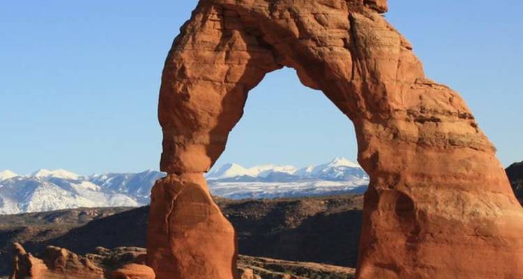 L'emblématique Delicate Arch se dresse contre un ciel bleu dégagé avec des chaînes de montagnes enneigées au-delà.