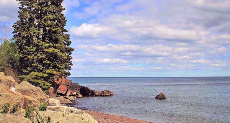 Rivage rocheux du lac Supérieur avec des sapins et un ciel partiellement nuageux s'étendant sur une eau bleue calme