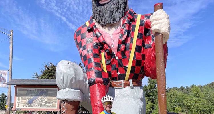 Un cycliste posant devant la statue géante colorée du bûcheron Paul Bunyan sur un ciel bleu dégagé