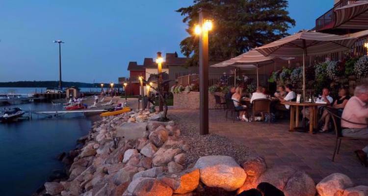 Terrasse de restaurant en bord de l'eau au crépuscule avec des clients assis sous des parasols à côté de petits bateaux au quai
