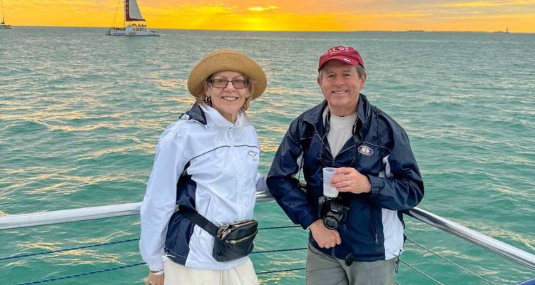 Glimlachend stel in lichte jasjes staat op een bootdek met een levendige oranje zonsondergang boven turquoise oceaanwater achter hen