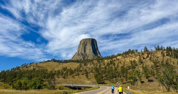 Fietsers rijden naar het dramatische monoliet van Devils Tower onder gestreepte blauwe hemels.