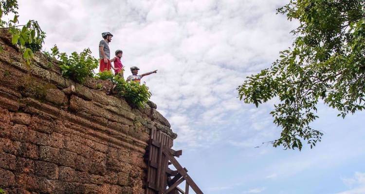 Cyclists stand atop an ancient laterite wall pointing across the Cambodian landscape under a dramatic sky