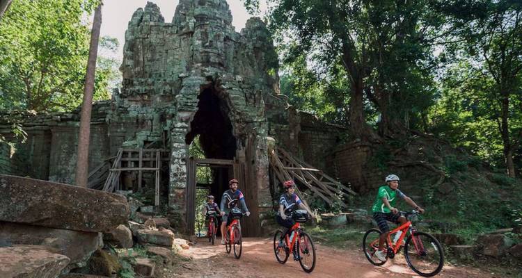 Cyclists ride red bikes through an ancient stone gate deep in the Angkor jungle.