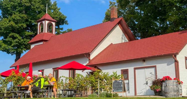 Fietsers ontspannen zich aan picknicktafels buiten een schilderachtig wit gebouw met rood dak en parasols, omgeven door wingerd.