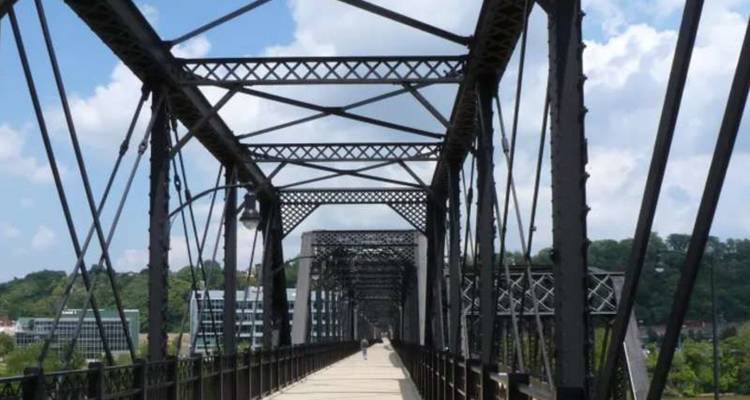 Voetgangers- en fietserbrug met stalen vakwerkconstructies die zich uitstrekt over de rivier in de richting van de stadshorizon op een zonnige dag.