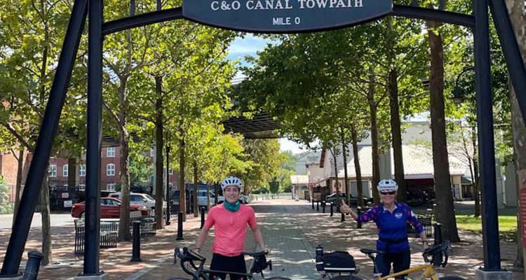 Twee fietsers staan met hun fietsen onder de zwarte metalen boog die de ingang van het C&O Canal Towpath-pad markeert, omgeven door bladrijke bomen en bakstenen paden.