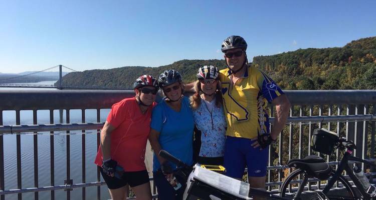 Vier fietsers poseren op een brug met een hangbrug in de verte en een rivier eronder