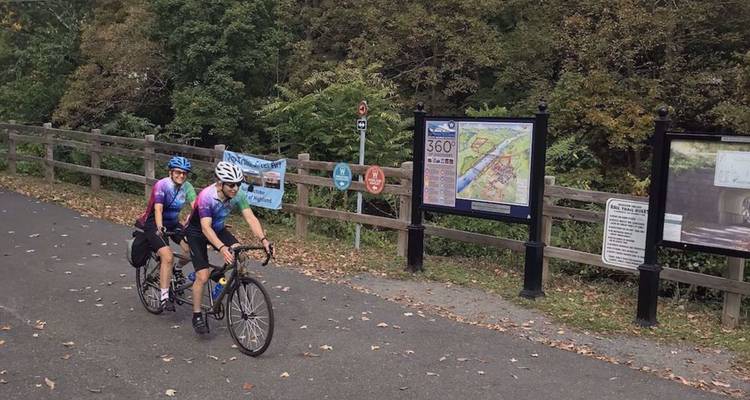 Twee fietsers rijden langs bewegwijzering en kaarten op een verhard pad dat wordt omzoomd door hekwerk