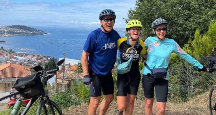 Three smiling cyclists pose on a hilltop overlooking a coastal town and blue Atlantic waters.