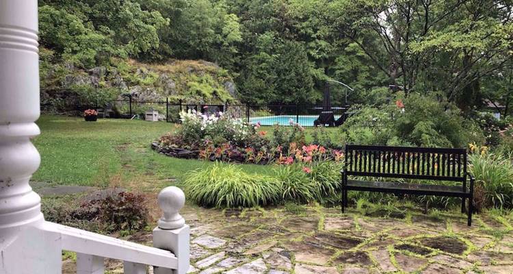 Jardin humide de pluie avec patio en pierre, parterres de fleurs, banc et une piscine distante entourée d'affleurements rocheux boisés.