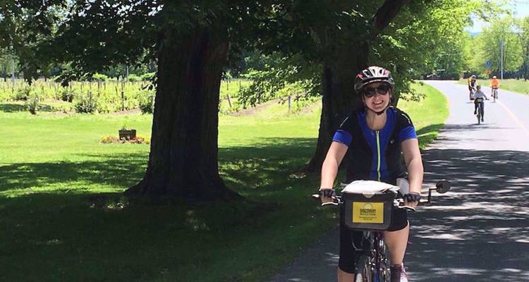 Une femme sourit en faisant du vélo le long d'une route rurale ombragée avec des vignobles et un autre cycliste au loin.