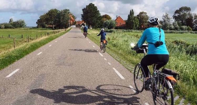Cyclists ride along a quiet rural road through Dutch farmland under partly cloudy skies.