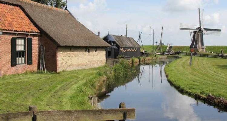 Rural Dutch scene with thatched barns, a narrow canal and distant windmill under soft clouds.