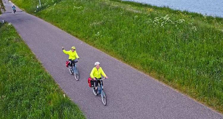 Two touring cyclists in bright jackets pedalling along a raised dyke path beside open water.