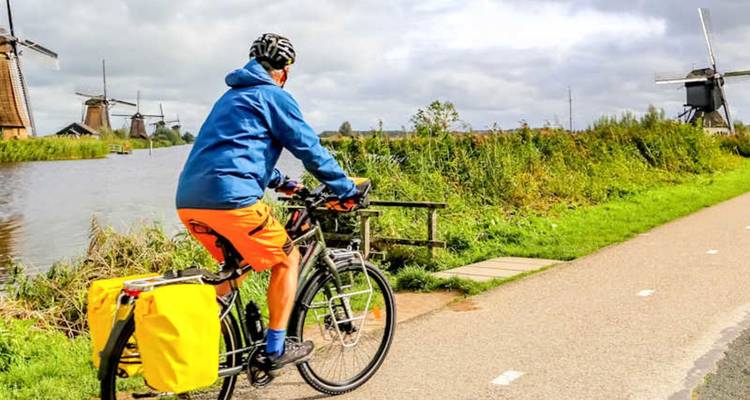 Touring cyclist on a paved path with iconic Dutch windmills lining a waterway.