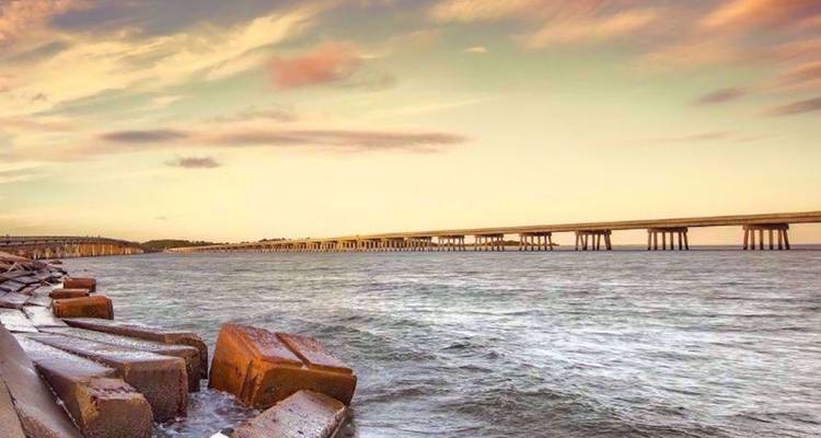 Lange betonnen brug die zich uitstrekt over kustwater onder pastelkleurige zonsonderganghemel.
