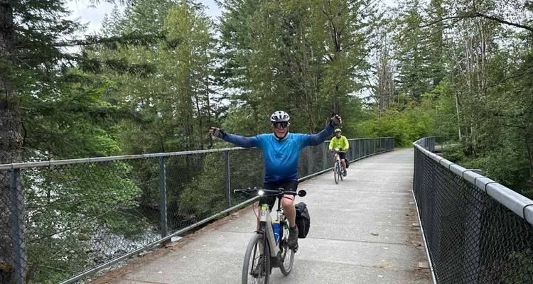 Les cyclistes traversent à vélo un pont suspendu dans les arbres, clôturé, qui traverse une dense forêt du Pacifique Nord-Ouest.
