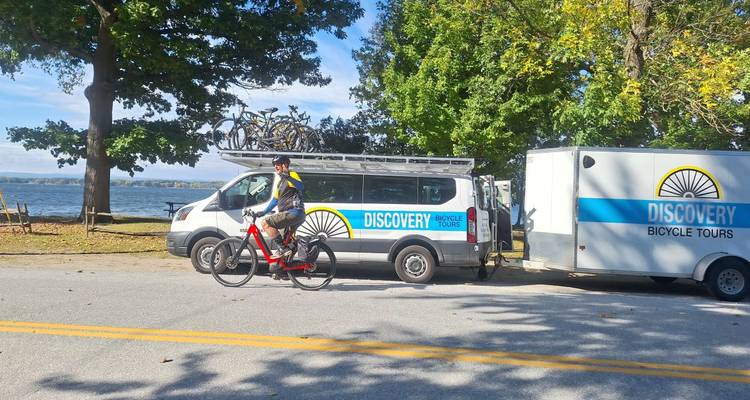 Fietser rijdt langs een Discovery Bicycle Tours busje en aanhanger met huisstijl die naast een meer geparkeerd staan
