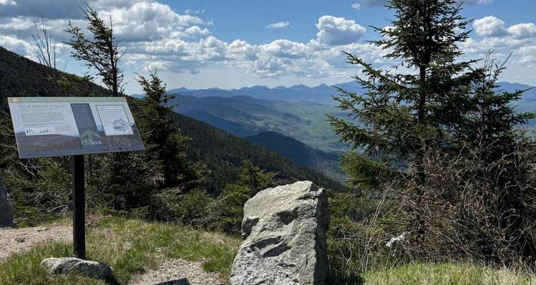 Panneau d'interprétation et rocher offrant une vue panoramique impressionnante sur les montagnes sous un ciel ensoleillé.