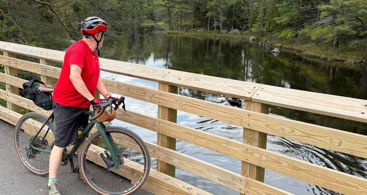 Cycliste en maillot rouge qui fait une pause sur un pont en bois au-dessus d'une rivière forestière aux reflets miroirs, observant la faune.