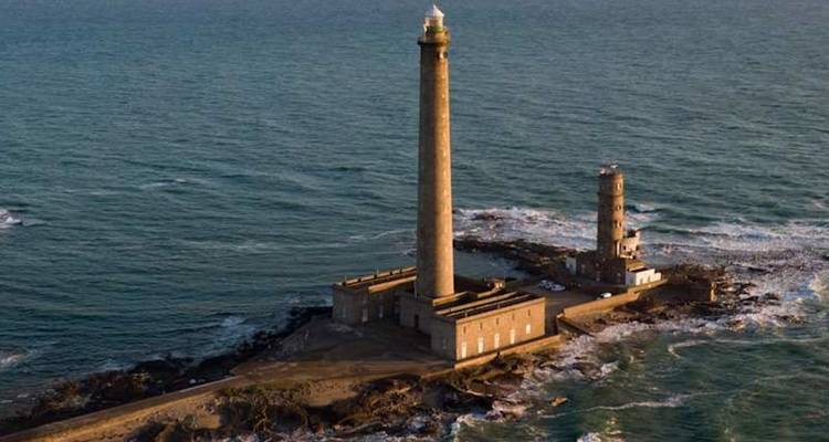 Vue aérienne d'un phare imposant s'élevant d'un îlot rocheux au milieu d'une mer bleu profond, lumière du soir.