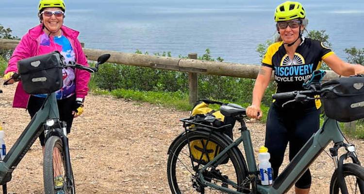 Two cyclists wearing bright helmets pose with their e-bikes on a coastal overlook.