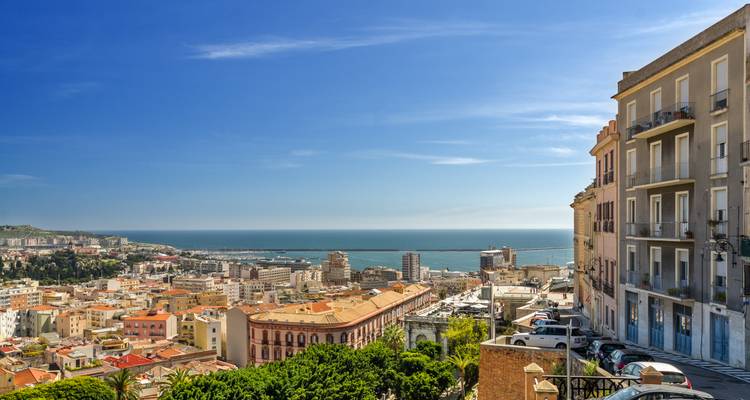 Vista elevada sobre los tejados de terracota de Cagliari hacia el mar Mediterráneo bajo un cielo despejado.