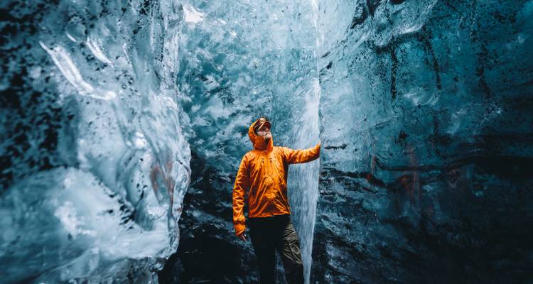 Adventurer in bright orange jacket exploring a dazzling blue ice cave with towering frozen walls.