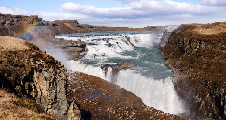 Powerful multi-tiered Gullfoss waterfall thundering through a rugged canyon with a faint rainbow in the mist.