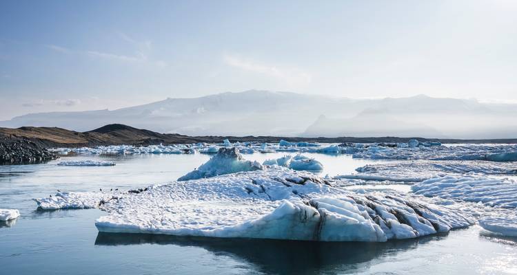 Calm blue waters dotted with icebergs at Jökulsárlón glacier lagoon under a hazy mountain backdrop.