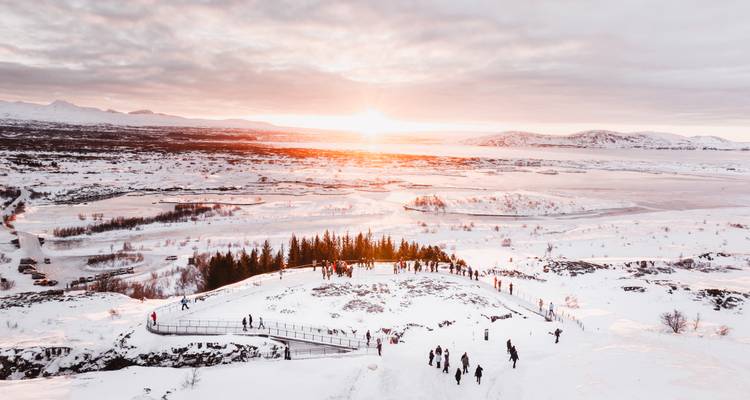 Snow-covered valley at sunset with tourists gathered on a ridge overlooking a frozen landscape.