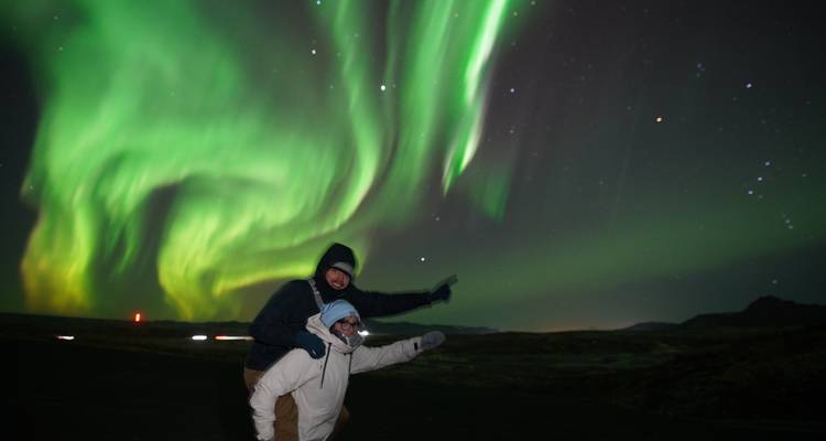 Couple posing playfully beneath vibrant green northern lights streaking across a starry sky.