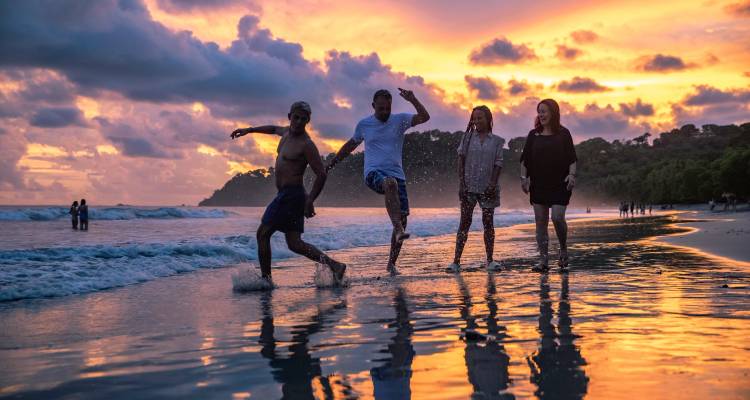 Friends splashing in shallow surf during a vivid orange and pink sunset.