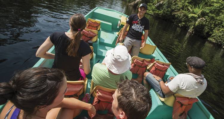 Travelers sit in an open boat navigating a narrow jungle waterway lined with lush greenery.