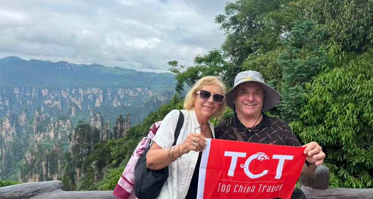 Una pareja sonriente sostiene una bandera de tour en un mirador pintoresco con montañas de pilares dramáticas y un bosque exuberante detrás de ellos.