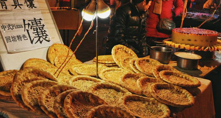 Puesto de comida en mercado callejero mostrando grandes panes planos redondos bajo lámparas cálidas con señalización china y clientes agrupados usando chaquetas de invierno.
