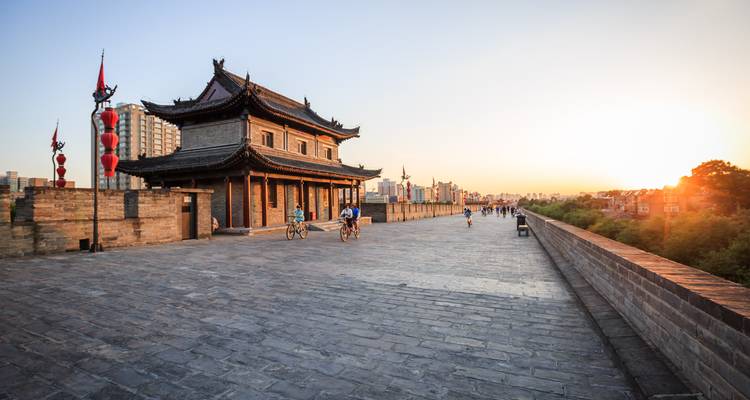 Los ciclistas recorren las antiguas murallas de piedra de Xi'an mientras el sol se pone, proyectando una luz dorada sobre los faroles rojos y las torres tradicionales.