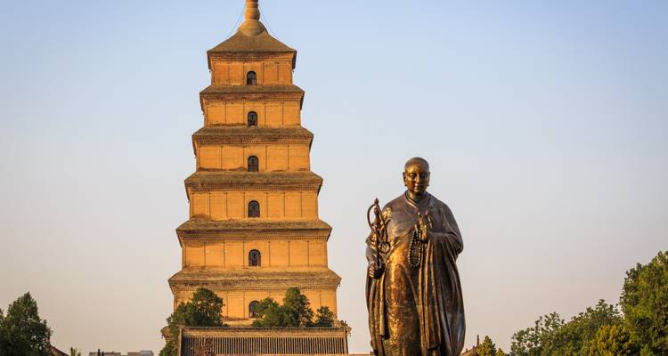 Estatua del monje Xuanzang de pie frente a la Gran Pagoda del Ganso Salvaje de varios pisos brillando en la cálida luz de la tarde contra un cielo despejado.