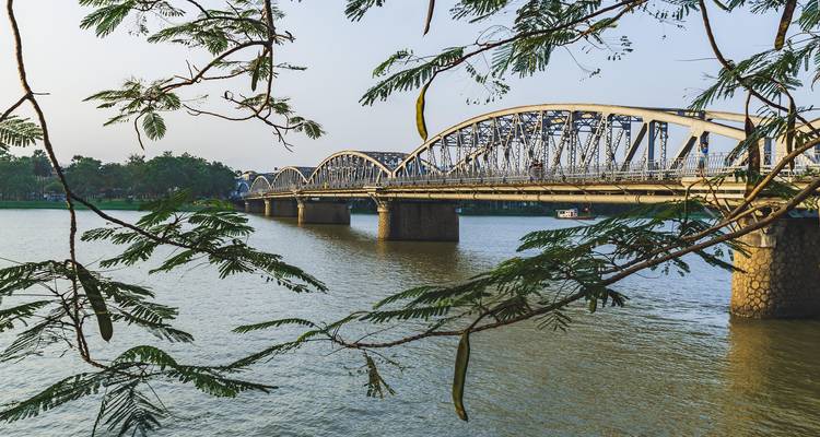 Pont en arc d'acier franchissant un large fleuve, encadré par des branches feuillues sur fond de ciel dégagé à Hue.