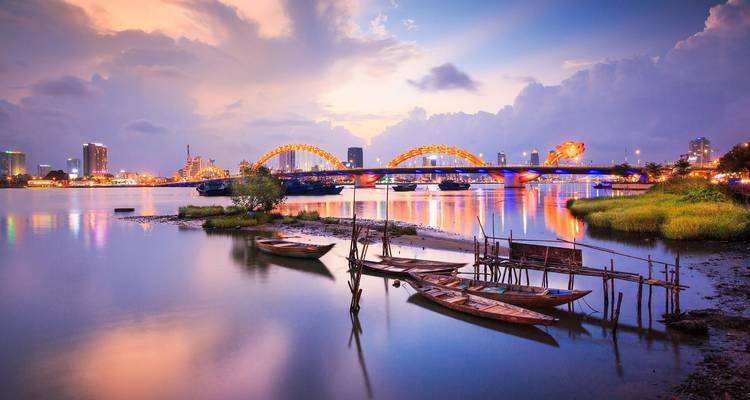 Panorama crépusculaire du pont du Dragon de Da Nang s'arquant au-dessus de la rivière miroir avec les lumières de la ville se reflétant dans l'eau calme.