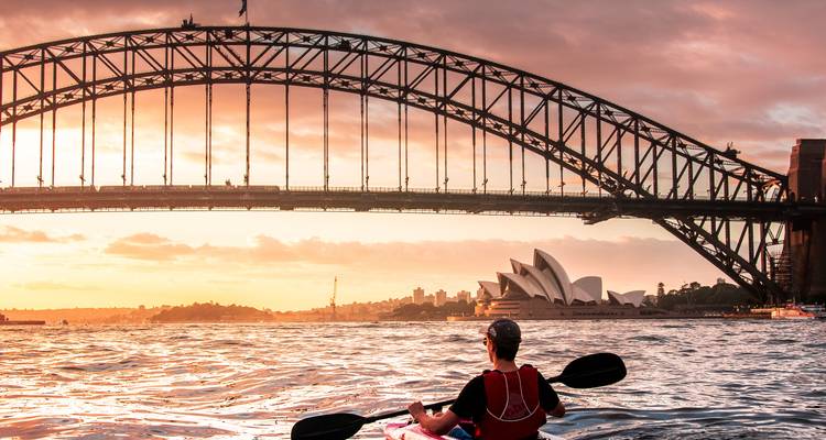 Kajakfahrer paddelt bei Sonnenuntergang über glitzerndes Wasser unter dem Bogen der Sydney Harbour Bridge mit dem Opernhaus dahinter