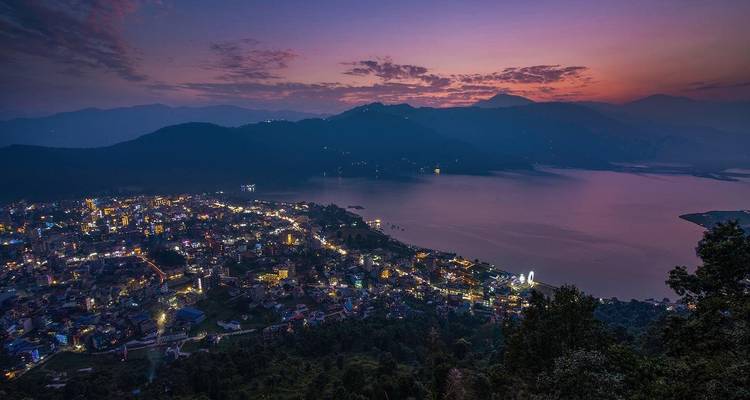Panorama del atardecer con las luces de la ciudad de Pokhara reflejándose en el lago Phewa con las montañas recortadas contra el cielo.